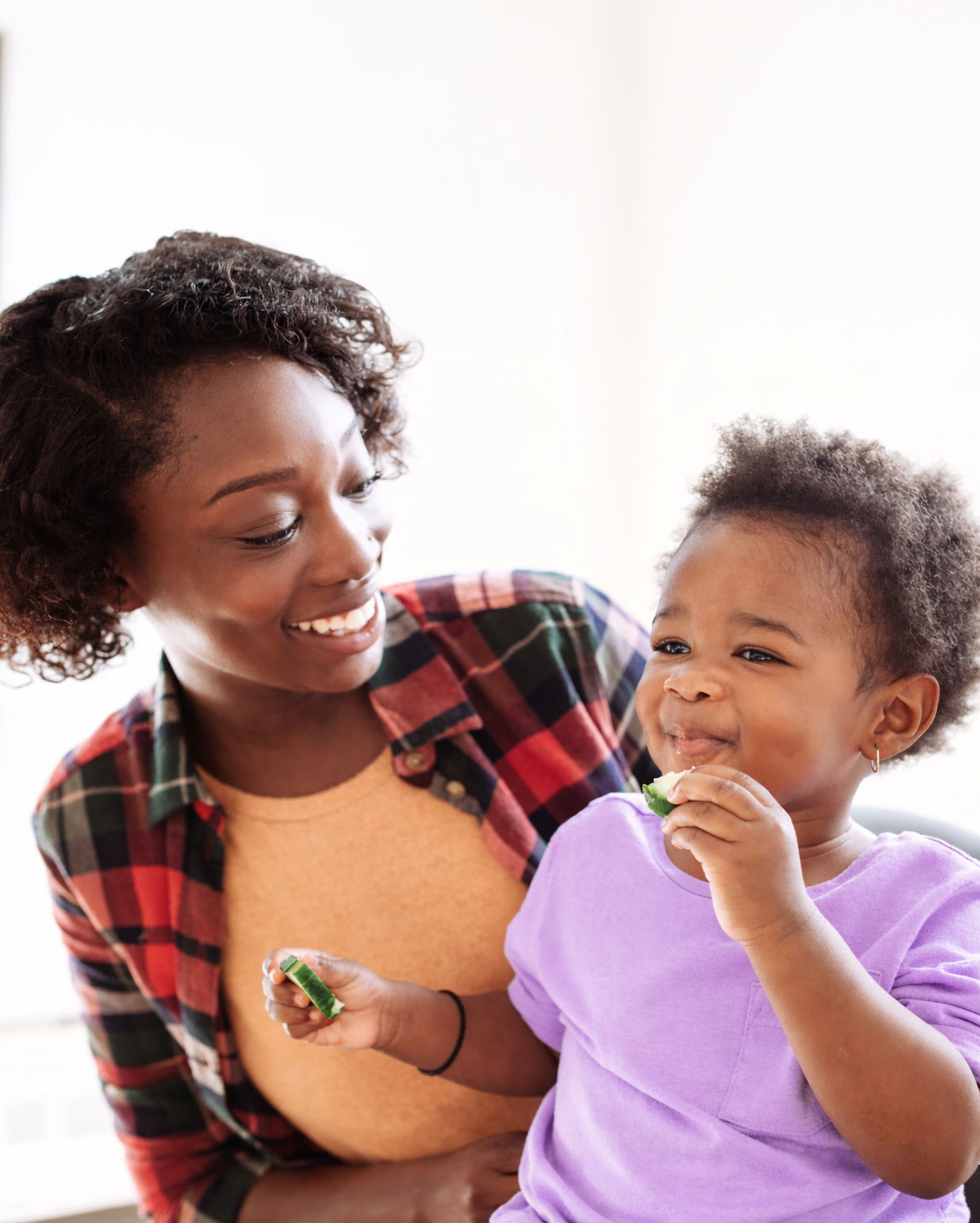 Child eating cucumber, smiling adult nearby