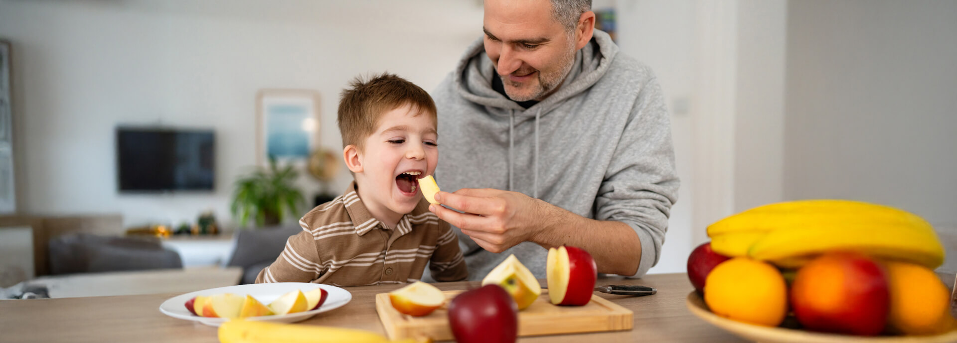 Man feeding child apple slice at kitchen table