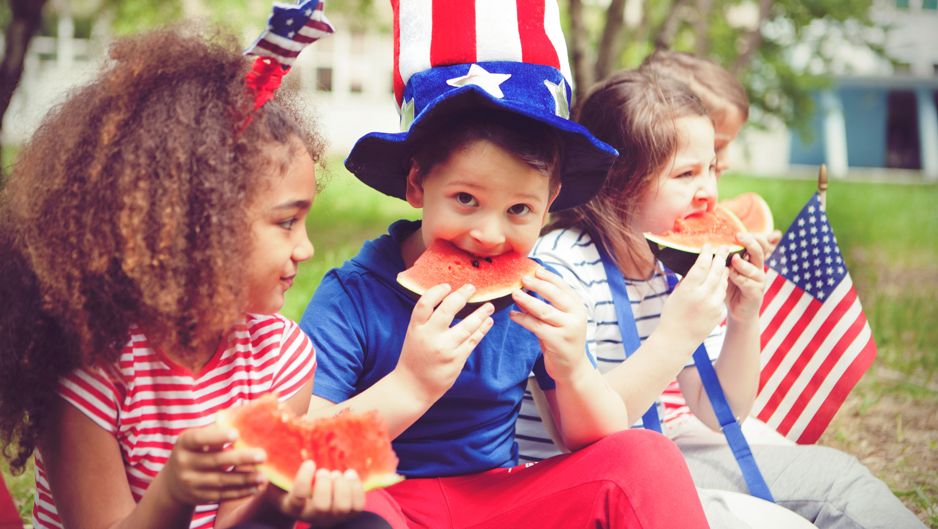 Children eating watermelon, wearing festive hats, flags nearby